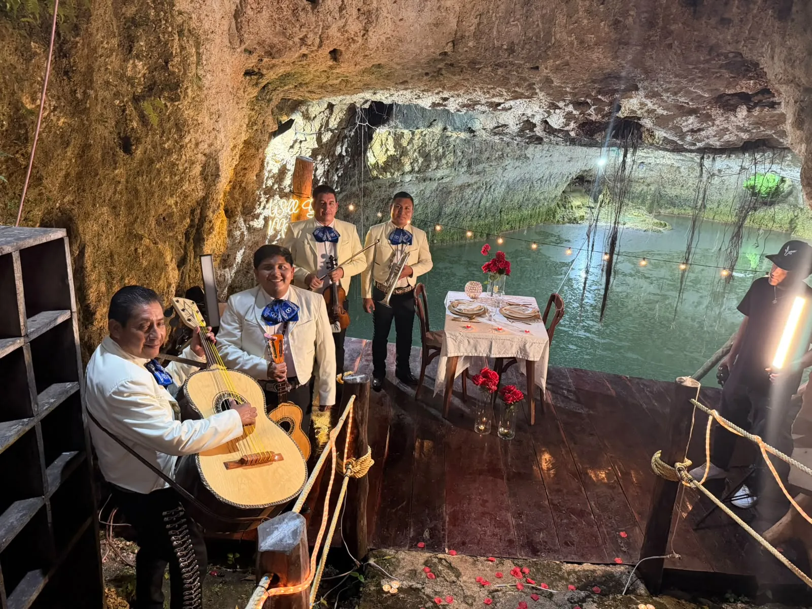 Mariachi con trajes claros y moños azules junto a una mesa de cena romántica para dos dentro de un cenote iluminado con luces cálidas