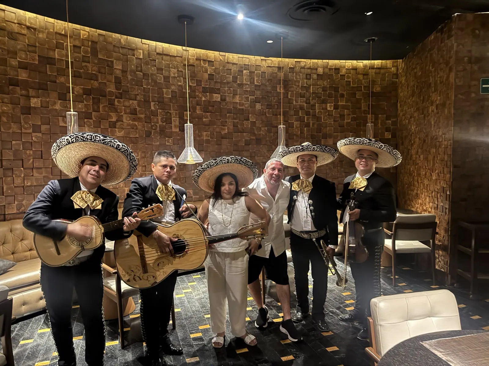 Mariachi con sombreros de palma y moños dorados posando con dos comensales en un restaurante de muros de madera