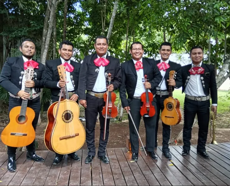 Seis músicos de mariachi con moños rosas y sus instrumentos sobre una terraza de madera rodeada de árboles
