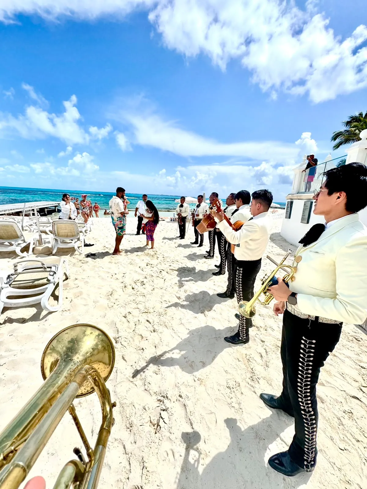 Mariachi tocando en la arena con el mar turquesa al fondo mientras una pareja se abraza, con una trompeta en primer plano